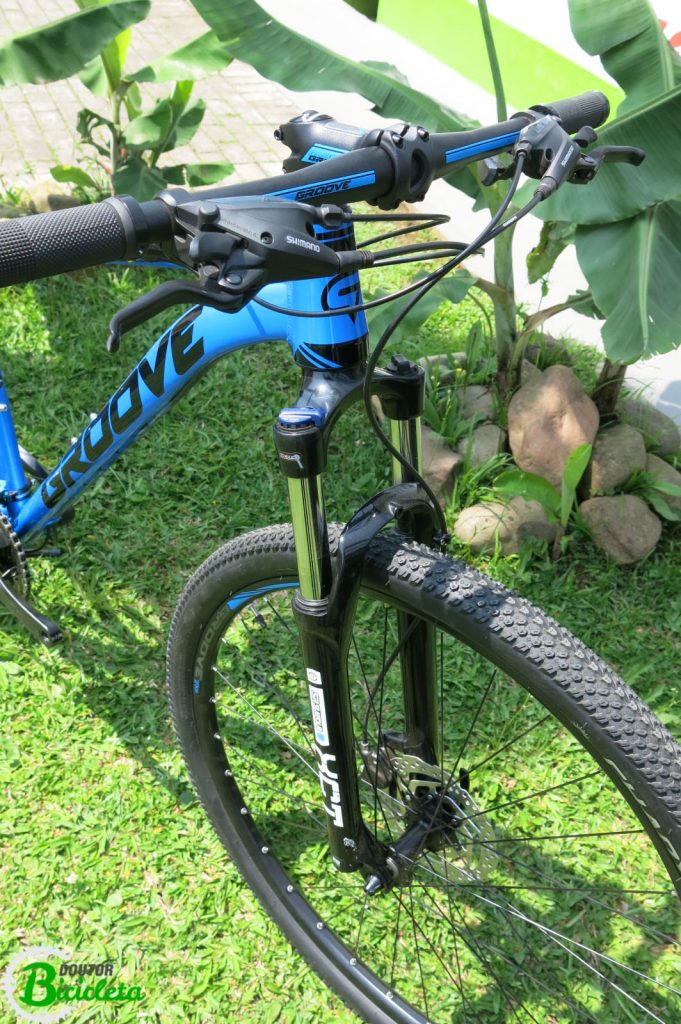 Suspensão dianteira SR Suntour XCT com trava, aros de 29", freios à disco hidráulicos, alavanca de câmbio com manete de freio integrado.